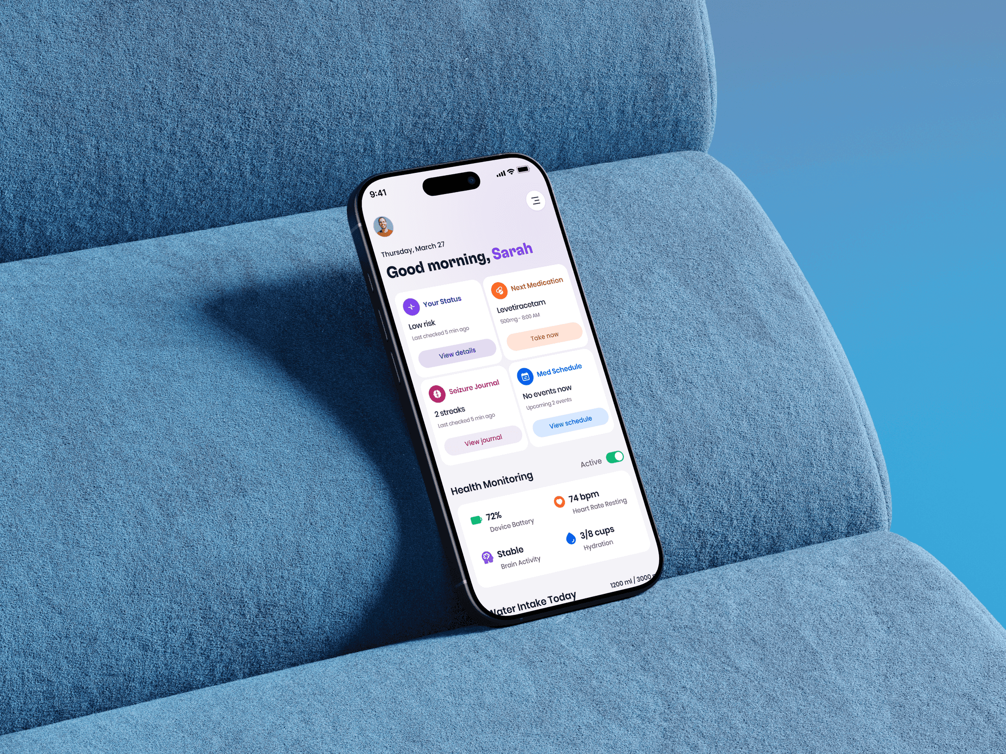 A phone leaning against the cushions of a blue couch, showing the redesigned home screen: a morning greeting, a low-risk status card, the next medication with a Take now button, seizure journal and medication schedule cards, and health monitoring readings for device battery, heart rate, brain activity and hydration.